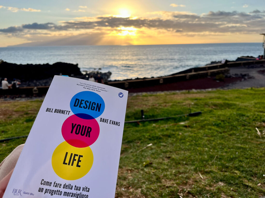Libro Design your lide, con sfondo su prato e in fondo oceano, al tramondo col sole dietro le nuvole. Foto di Erica Bortolussi